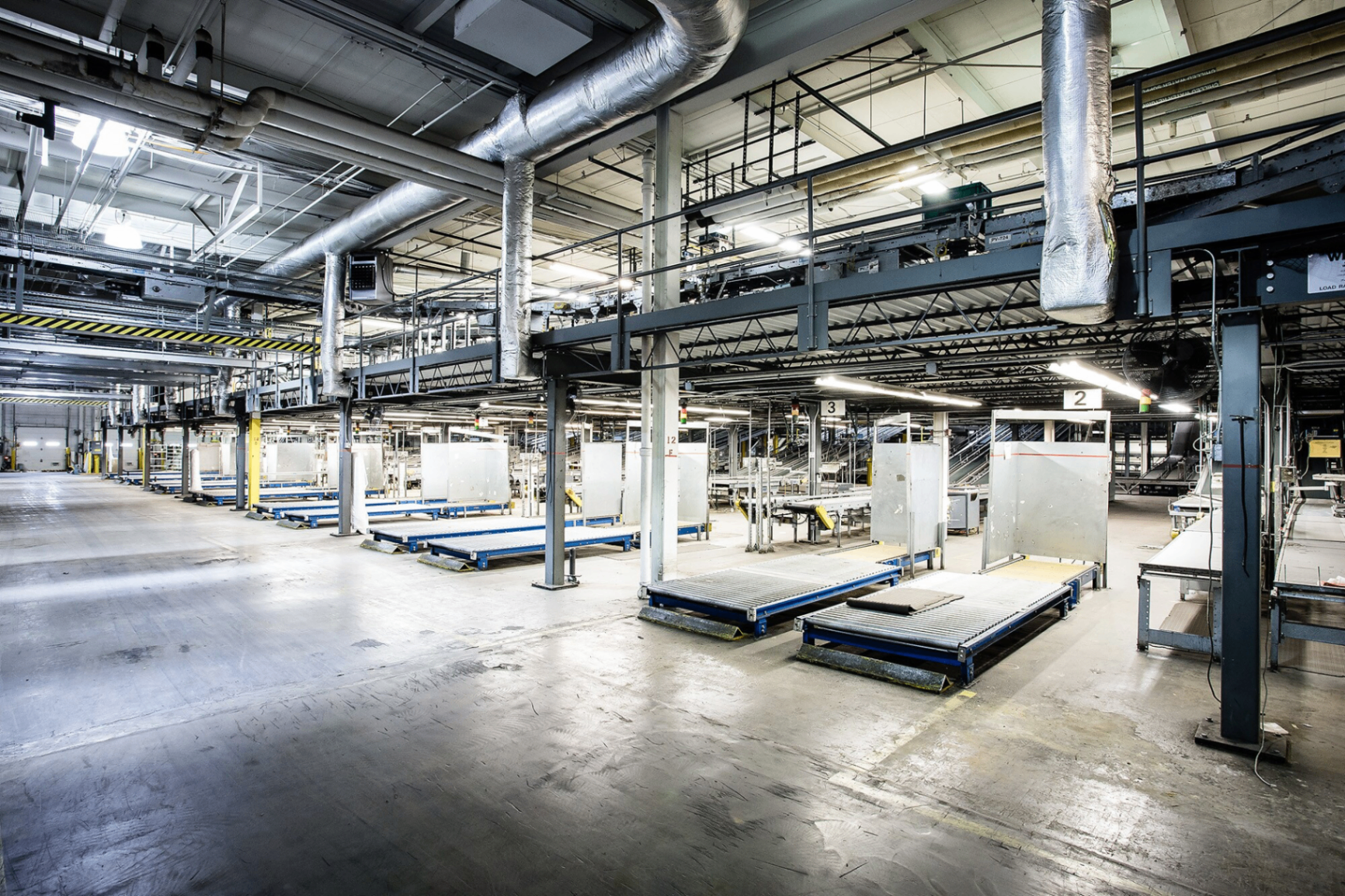 Interior of a large industrial manufacturing hall with conveyor systems, steel beams, and loading docks